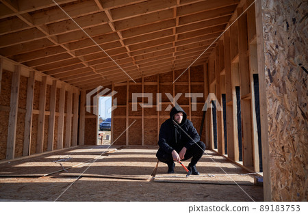 Male engineer building wooden frame house in the Scandinavian style barnhouse. Man developer on construction site, inspecting quality of work on sunny day, holding hammer in hands. 89183753