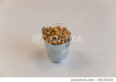 A full metal bucket of dry pelleted dog food. Small silver thigh with dog food against a gray background. Pet supplies. Selective Focus. A full metal bucket of dry pelleted dog food. Small silver thigh with dog food against a gray background. Pet supplies. Selective Focus. 89184288