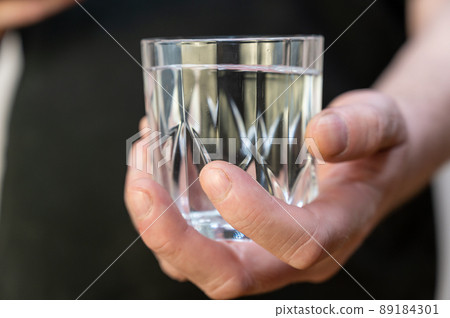 A close-up of a hand holding a glass of water. A mature man in a black shirt holds a glass of clear drinking water in his hand. 89184301