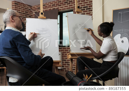 Senior man teacher supervising young student during drawing class in creativity studio developing artistic skill. Artist sketching vase model on canvas using draw technique. 89187355