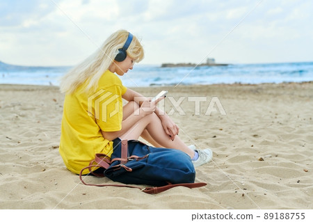 Teenage female with backpack in headphones using smartphone, on sandy beach 89188755