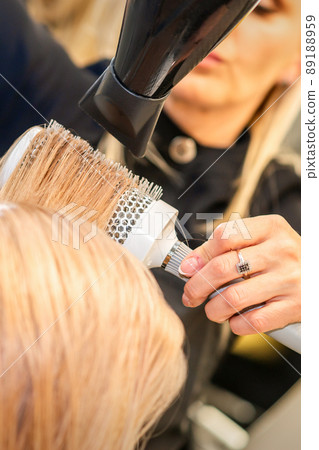Drying straight blond hair with black hairdryer and white round brush in hairdresser salon, close up Drying straight blond hair with black hairdryer and white round brush in hairdresser salon, close up 89188959