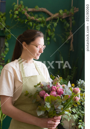 Businesswoman. Young woman florist in an apron and glasses checks a floral bouquet. Beautiful flowers to order. The concept of young entrepreneurs Businesswoman. Young woman florist in an apron and glasses checks a floral bouquet. Beautiful flowers to order. The concept of young entrepreneurs 89190060