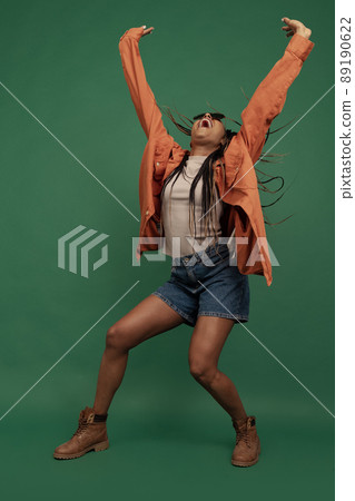 Emotional african girl in shorts and shirt dancing, having fun isolated on dark green background. Concept of beauty, art, fashion, youth and emotions 89190622