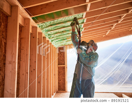 Male builder insulating wooden frame house. Man worker spraying polyurethane foam inside of future cottage, using plural component gun. Construction and insulation concept. Male builder insulating wooden frame house. Man worker spraying polyurethane foam inside of future cottage, using plural component gun. Construction and insulation concept. 89191521