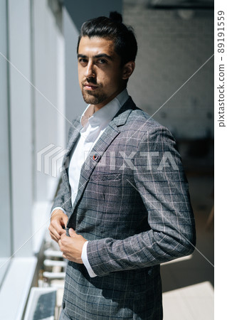 Vertical portrait of stylish handsome business man wearing fashion suit standing near window in modern office room, looking at camera. Front view of bearded businessman posing at workplace. Vertical portrait of stylish handsome business man wearing fashion suit standing near window in modern office room, looking at camera. Front view of bearded businessman posing at workplace. 89191559