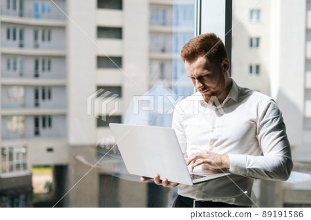 Medium shot portrait of focused young man working typing on laptop holding with one hand standing in modern office room near window, in sunny summer day on background of buildings. Medium shot portrait of focused young man working typing on laptop holding with one hand standing in modern office room near window, in sunny summer day on background of buildings. 89191586
