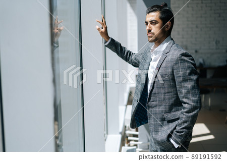 Portrait of thinking elegant business man wearing fashion suit standing in modern office room, looking out window. Front view of serious bearded businessman posing at workplace. 89191592