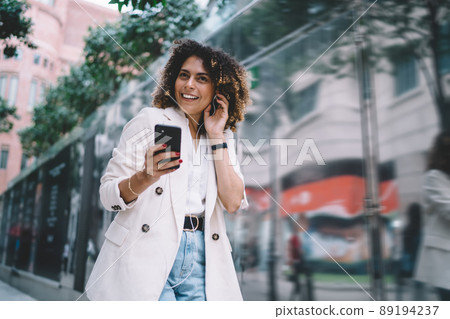 Joyful female generation Z with mobile phone in hand smiling at streets 89194237