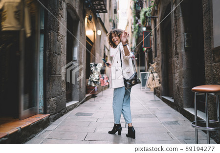 Trendy dressed Caucaisan hipster girl with mobile device in hand waving at touristic street Trendy dressed Caucaisan hipster girl with mobile device in hand waving at touristic street 89194277