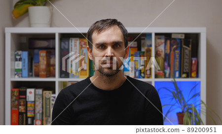 A handsome middle-aged Caucasian man in a black sweatshirt in a room against a rack of board games. A handsome middle-aged Caucasian man in a black sweatshirt in a room against a rack of board games. 89200951