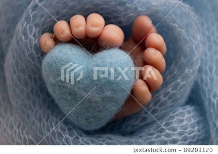 The tiny foot of a newborn baby. Soft feet of a new born in a purple, lilac wool blanket. Close up of toes, heels and feet of a newborn. Knitted purple, lilac heart in the legs of a baby.  89201120