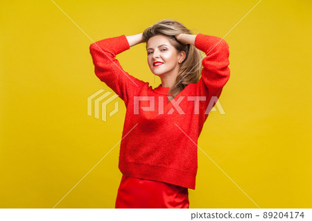 Portrait of impressive happy blonde woman with red lipstick in bright sweater standing with hands in hair, looking at camera with seductive smile. indoor studio shot isolated on yellow background 89204174