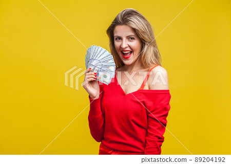 Portrait of amazed wealthy young woman in red dress standing, holding dollar bills, looking at camera with open mouth, surprised with lottery winning. indoor studio shot isolated on yellow background 89204192