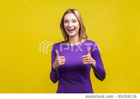 Good job. Portrait of extremely happy woman in tight purple dress showing thumbs up and smiling broadly at camera, overjoyed excited expression. indoor studio shot isolated on yellow background Good job. Portrait of extremely happy woman in tight purple dress showing thumbs up and smiling broadly at camera, overjoyed excited expression. indoor studio shot isolated on yellow background 89204216