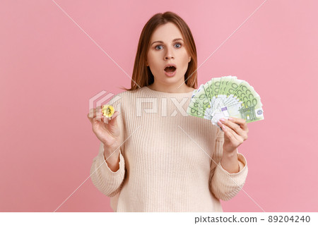 Astonished surprised woman holding gold coin of crypto currency and big fan of euro banknotes, looking at camera with open mouth, wearing white sweater. Indoor studio shot isolated on pink background. Astonished surprised woman holding gold coin of crypto currency and big fan of euro banknotes, looking at camera with open mouth, wearing white sweater. Indoor studio shot isolated on pink background. 89204240