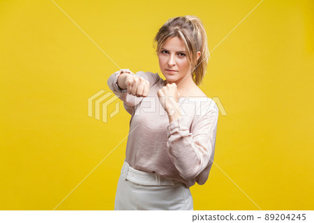 Portrait of brave confident young woman with blonde hair in casual beige blouse standing, holding clenched fists up ready to boxing, fighting spirit. indoor studio shot, isolated on yellow background 89204245