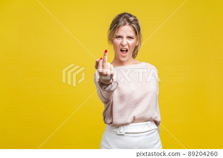 Portrait of angry dissatisfied young woman with fair hair in casual beige blouse standing, showing middle finger f you, looking at camera with hate. indoor studio shot isolated on yellow background 89204260
