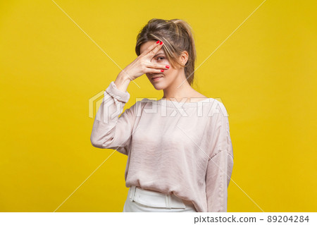 Portrait of curious beautiful young woman with fair hair in casual beige blouse standing, peeking through fingers with suspicious and prying eyes. indoor studio shot isolated on yellow background 89204284