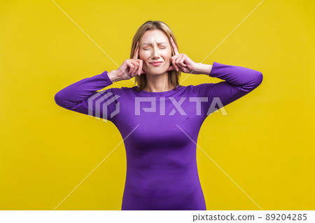 Intense headache. Portrait of upset sick woman in elegant tight purple dress standing touching her temples and grimacing from pain, suffering migraine. indoor studio shot isolated on yellow background 89204285