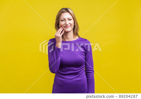 Toothache. Portrait of sick unhappy woman in elegant tight purple dress suffering sensitive teeth, grimacing from acute dental pain, looking at camera. indoor studio shot isolated on yellow background Toothache. Portrait of sick unhappy woman in elegant tight purple dress suffering sensitive teeth, grimacing from acute dental pain, looking at camera. indoor studio shot isolated on yellow background 89204287