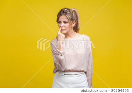 You are liar. Portrait of dishonest young woman with fair hair in casual beige blouse standing, touching her nose with finger, showing lie gesture. indoor studio shot isolated on yellow background 89204305