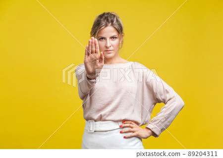 Portrait of confident serious young woman with fair hair in casual beige blouse standing, looking at camera, showing stop gesture, warning expression. indoor studio shot isolated on yellow background Portrait of confident serious young woman with fair hair in casual beige blouse standing, looking at camera, showing stop gesture, warning expression. indoor studio shot isolated on yellow background 89204311