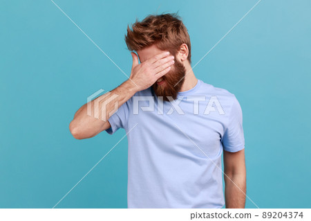 Don't want to look at this. Portrait of bearded man covering eyes and turning with disgust from something shameful, scared afraid to see. Indoor studio shot isolated on blue background. 89204374