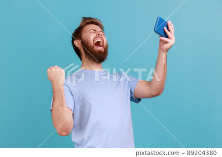 Portrait of extremely happy handsome bearded man gamer playing video game on mobile phone, celebrating victory, complete level. Indoor studio shot isolated on blue background. 89204380