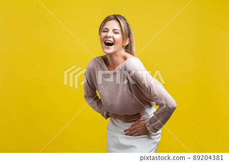Portrait of happy cute woman with blonde hair in casual beige blouse standing, holding her belly and laughing out loud, looking at camera with joy. indoor studio shot isolated on yellow background 89204381