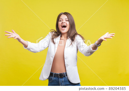 Portrait of delighted beautiful brunette woman in jacket and jeans raising hands in welcome gesture screaming for joy, excited to see you and going to hug. studio shot isolated on yellow background 89204385