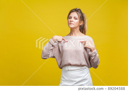Portrait of arrogant proud woman with fair hair in casual blouse standing, pointing at herself, boasting personal achievements at camera. indoor studio shot isolated on yellow background 89204388