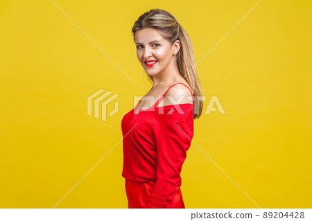 Half turn portrait of happy elegant young lady with neat ponytail hairstyle, bright makeup in red dress standing, looking at camera with toothy smile. indoor studio shot isolated on yellow background Half turn portrait of happy elegant young lady with neat ponytail hairstyle, bright makeup in red dress standing, looking at camera with toothy smile. indoor studio shot isolated on yellow background 89204428