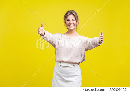 Come into my arms. Portrait of friendly beautiful woman with fair hair in casual beige blouse standing with stretching hands, wants to hug tight. indoor studio shot isolated on yellow background Come into my arms. Portrait of friendly beautiful woman with fair hair in casual beige blouse standing with stretching hands, wants to hug tight. indoor studio shot isolated on yellow background 89204432