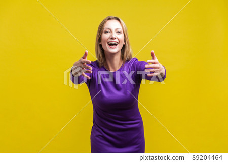 Come into my arms, free hug. Portrait of adorable hospitable woman in elegant tight purple dress smiling and reaching out hands, going to embrace. indoor studio shot isolated on yellow background 89204464