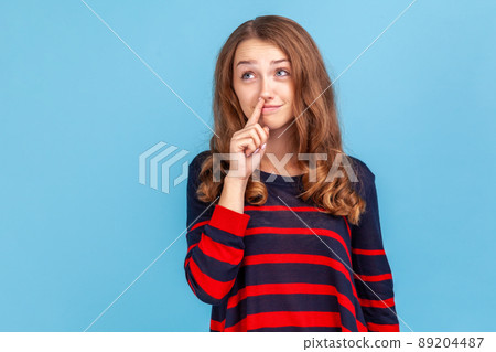 Childish woman wearing striped casual style sweater holding finger in her nose, uncultured bored girl having fun, bad manners, looking away. Indoor studio shot isolated on blue background. Childish woman wearing striped casual style sweater holding finger in her nose, uncultured bored girl having fun, bad manners, looking away. Indoor studio shot isolated on blue background. 89204487