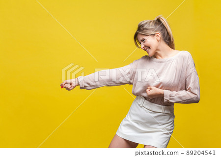 Profile side view of positive assertive young woman with fair hair in casual beige blouse standing with pulling gesture, much effort face expression. indoor studio shot isolated on yellow background 89204541