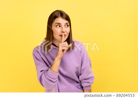 Shh, it's big secret. Beautiful cunning woman smiling, showing gesture secret sign with finger near her lips, looking away, wearing purple hoodie. Indoor studio shot isolated on yellow background. Shh, it's big secret. Beautiful cunning woman smiling, showing gesture secret sign with finger near her lips, looking away, wearing purple hoodie. Indoor studio shot isolated on yellow background. 89204553