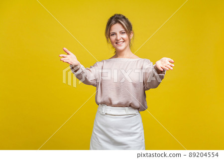 Portrait of happy generous young woman with fair hair in casual blouse standing with raised hands and looking at camera, welcoming or giving smth. indoor studio shot isolated on yellow background 89204554