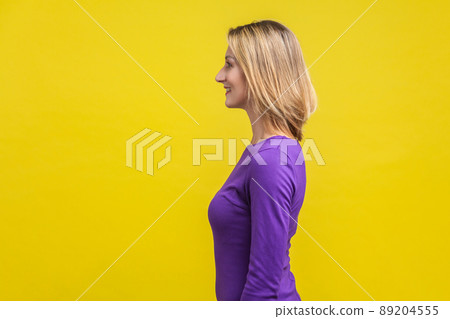 Side view of positive successful woman in purple dress standing with toothy smile, looking happy and content, empty copy space for advertisement . indoor studio shot isolated on yellow background 89204555