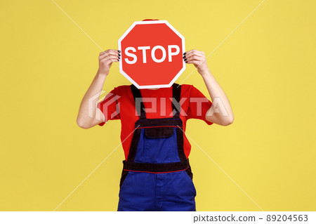 Portrait of worker woman standing hiding her face behind red stop sign, prohibitions and restrictions, wearing overalls and red cap. Indoor studio shot isolated on yellow background. 89204563