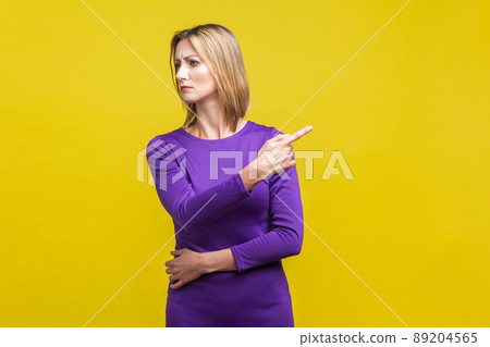 Portrait of angry bossy woman in elegant tight purple dress looking aside and pointing finger another way, asking to leave, dissatisfied expression. indoor studio shot isolated on yellow 89204565