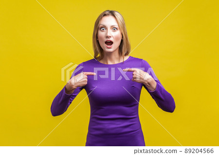Portrait of amazed woman in elegant purple dress looking at camera with open mouth and pointing at herself, shocked about own success. indoor studio shot isolated on yellow background 89204566