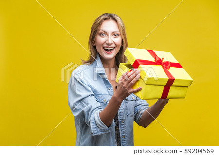 Portrait of joyous beautiful woman in denim shirt standing with opened gift box, looking at camera with toothy smile, satisfied with holiday present. studio shot isolated on yellow background 89204569