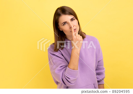 Portrait of woman looking with incredulous suspicious gaze and touching nose, gesturing you are liar, suspecting falsehood, wearing purple hoodie. Indoor studio shot isolated on yellow background. 89204595