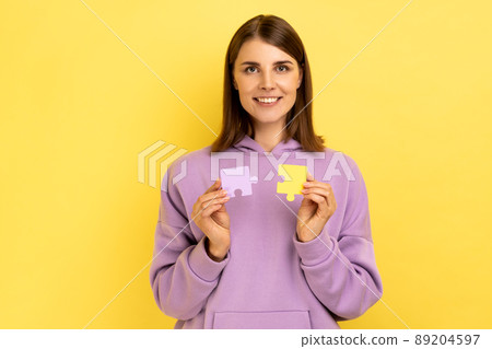 Smiling woman holding and showing two jigsaw puzzle pieces, matching and connecting puzzles, solving problems with good result, wearing purple hoodie. Indoor studio shot isolated on yellow background. Smiling woman holding and showing two jigsaw puzzle pieces, matching and connecting puzzles, solving problems with good result, wearing purple hoodie. Indoor studio shot isolated on yellow background. 89204597