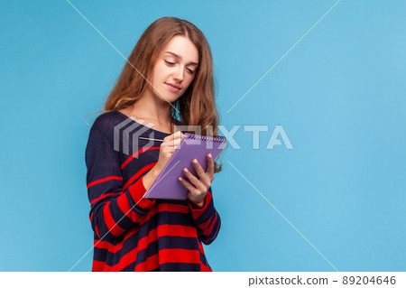 Positive woman wearing striped casual style sweater, writing in organizer, making to do list, designer drawing sketch or taking notes. Indoor studio shot isolated on blue background. 89204646