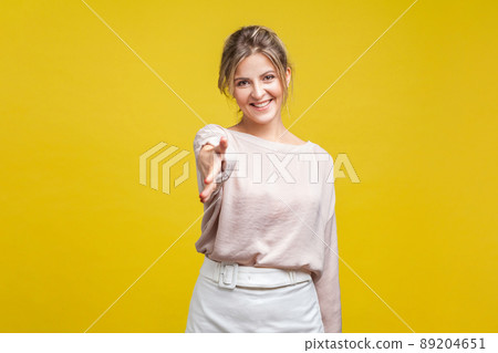 Portrait of friendly pretty woman with fair hair in casual beige blouse standing, looking at camera and offering hand for handshake, business greeting. indoor studio shot isolated on yellow background Portrait of friendly pretty woman with fair hair in casual beige blouse standing, looking at camera and offering hand for handshake, business greeting. indoor studio shot isolated on yellow background 89204651