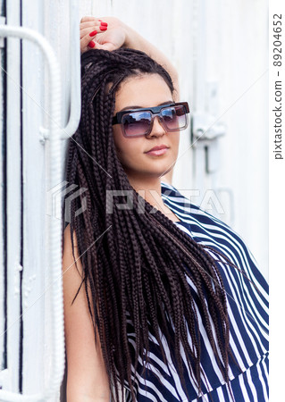 Portrait of young brunette woman with black afro american dreadlocks hairstyle in striped dress and sunglasses, standing, posing and looking at camera. indoor studio shot on white wall. Portrait of young brunette woman with black afro american dreadlocks hairstyle in striped dress and sunglasses, standing, posing and looking at camera. indoor studio shot on white wall. 89204652
