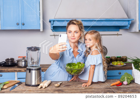 Charming woman and little girl taking selfie in modern kitchen, holding bowl of vegetable salad and posting photo in social network using cell phone, food blog about healthy nutrition, vegetarian diet Charming woman and little girl taking selfie in modern kitchen, holding bowl of vegetable salad and posting photo in social network using cell phone, food blog about healthy nutrition, vegetarian diet 89204653